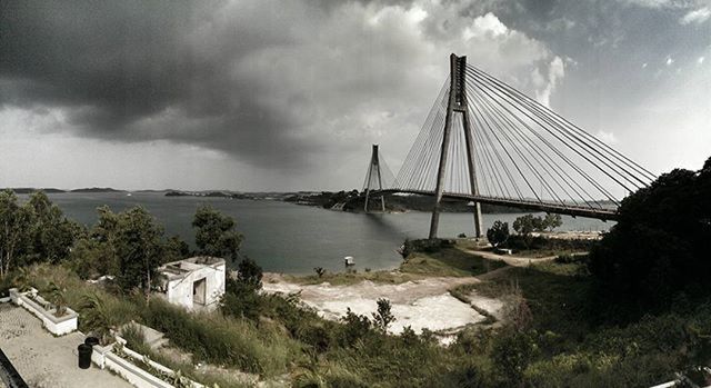 architecture, built structure, sky, bridge - man made structure, connection, transportation, cloud - sky, river, water, cloudy, engineering, building exterior, suspension bridge, city, cloud, bridge, tree, mode of transport, day, outdoors