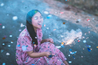 Cheerful woman holding illuminated sparkler while sitting outdoors