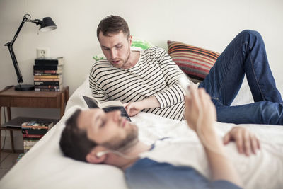 Young gay man reading book with partner listening music on bed at home