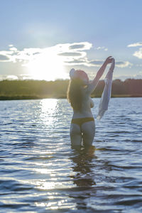 Rear view of topless young woman standing in lake against blue sky during sunset