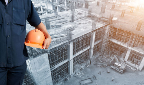 Midsection of construction worker holding hardhat at incomplete building