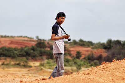 Young man with camera standing on hill against sky