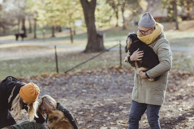 Smiling man carrying dog while walking towards women and pets at park