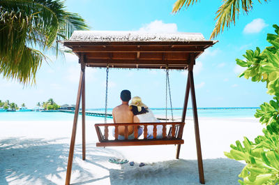 Couple sitting on swing at beach against sky