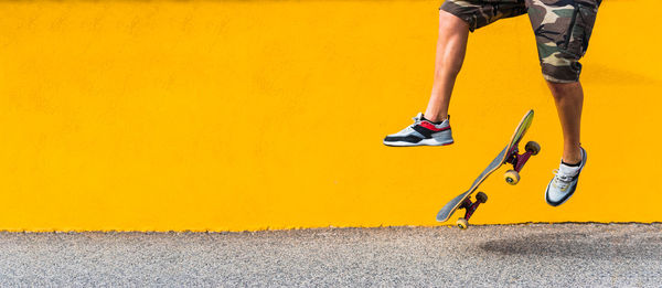 View of man on shorts legs jumping with skateboard on yellow background.