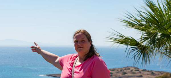 Portrait of mature woman pointing while standing against sea during sunny day