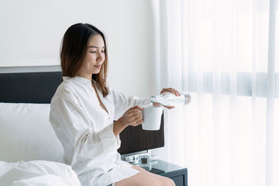 Smiling woman drinking water while sitting on bed at home