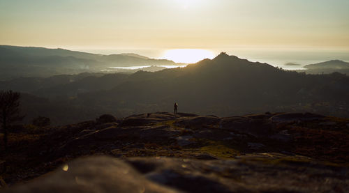 Distant view of woman standing against mountain and sky during sunset