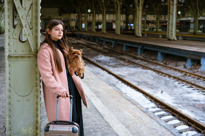 Beautiful young casual tourist woman with dog and suitcase waiting for train