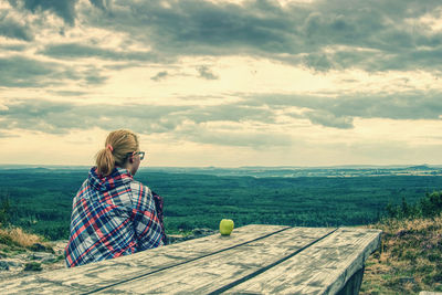 Blonde girl sits alone at table on relax place close edge abyss in mountains. apple on wooden desk 