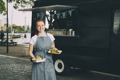 Portrait of smiling female owner holding indian food plate against commercial land vehicle