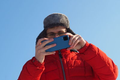 A young man of european appearance in a red jacket and a winter hat takes pictures from the phone in