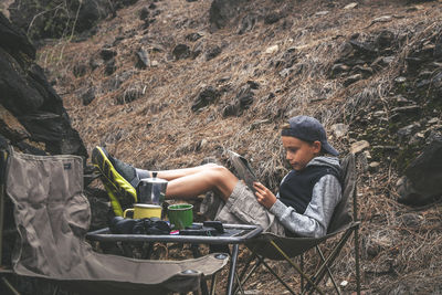 Full length of boy reading map while relaxing on chair at forest