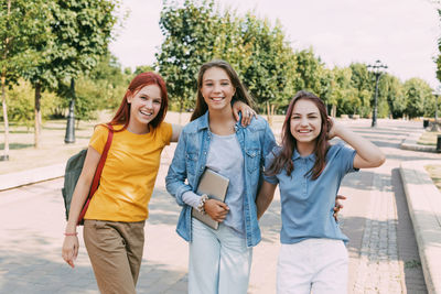 Three teenage girls are walking with a tablet and a backpack along the alley of the park 