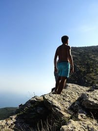 Rear view of shirtless man standing on rock against clear sky