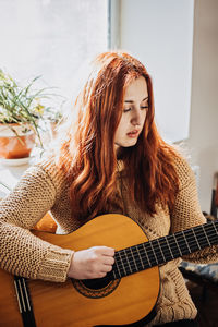 Unaltered candid portrait of young red haired woman in sweater playing acoustic guitar sitting by