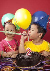 Boy blowing party popper while celebrating sister birthday against red background