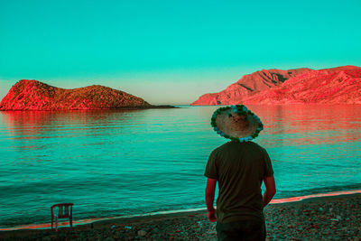 Rear view of man wearing hat standing at beach against clear blue sky during sunset