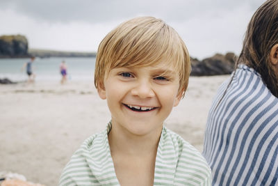 Portrait of smiling blond boy wrapped in towel by sister at beach