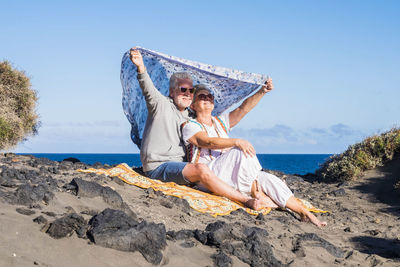 Senior couple sitting with sarong at beach against sky