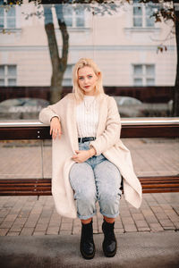 Cute young woman looking at camera while sitting on bus stop in city