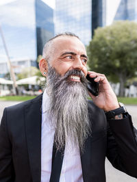 Confident smiling bearded male in classy suit having phone conversation while standing near road on street with modern buildings in city