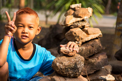 Portrait of boy gesturing peace sign while stacking rocks outdoors