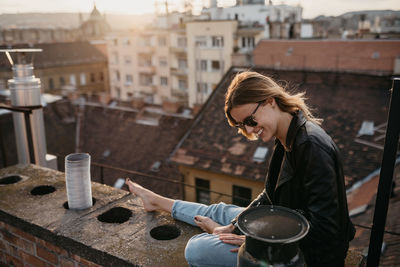 Happy woman wearing sunglasses while sitting on retaining wall in city