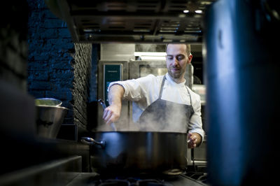 Young man in grey apron stirring big metal pan on restaurant kitchen