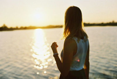 Rear view of woman standing by lake against sky during sunset