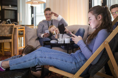 Teenage girl using smart phone while relaxing with family in living room at home