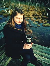 Woman in warm clothing holding bottle while sitting on bench by marsh