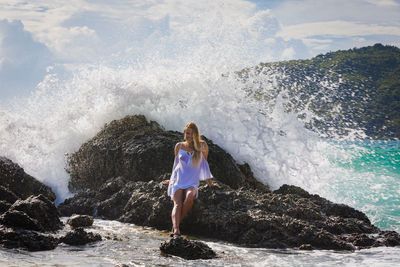 A young girl with long hair sits on a stone by the sea.