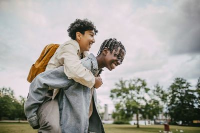 Side view of happy braided hair teenage boy piggybacking male friend at park