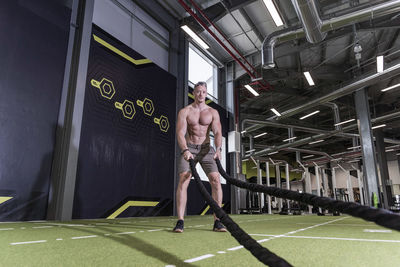 Portrait of shirtless man doing battle rope exercise at gym