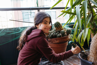 Sitting girl with pony tail wearing brown top taking care of plants in pot at home balcony