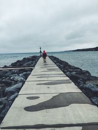 Rear view of man walking on pier over sea against cloudy sky