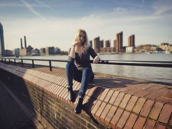 Portrait of young woman sitting on retaining wall by river against sky in city during sunny day