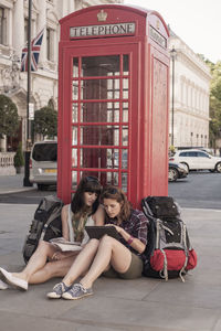 Female backpackers sitting with digital tablet against red telephone box in city