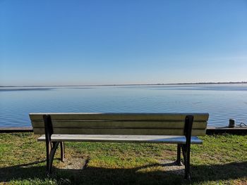 Empty bench by sea against clear blue sky