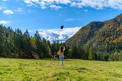 Rear view of young woman stading on green pasture, throwing hat in air.