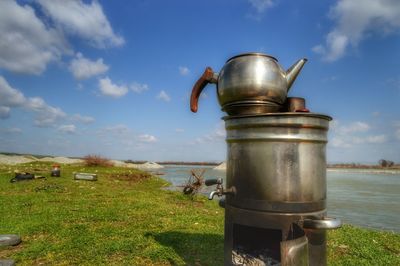 Teapot on metal container at riverbank against sky