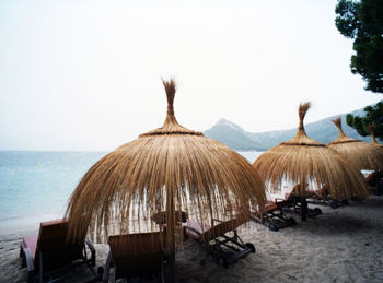 Lounge chairs by thatched roof parasols at beach against sky