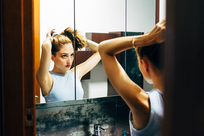 Young female in casual outfit standing in front of mirror and doing ponytail during daily routine in bathroom at home