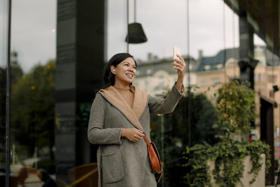 Smiling businesswoman on video call using smart phone while standing against glass building