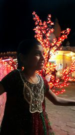 Rear view of smiling girl standing against illuminated christmas tree at night