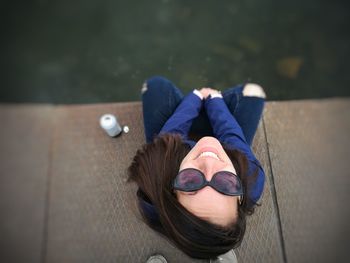 High angle portrait of smiling young woman in sunglasses sitting on jetty over lake