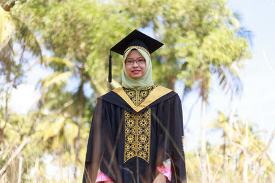 Portrait of young woman wearing hijab and graduation gown standing at park