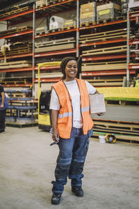 Full length of smiling female carpenter carrying cardboard box in warehouse