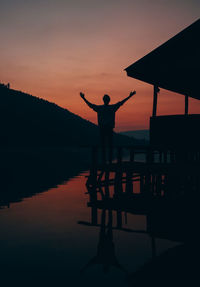 Silhouette man standing while hands raised by lake against sky during sunset